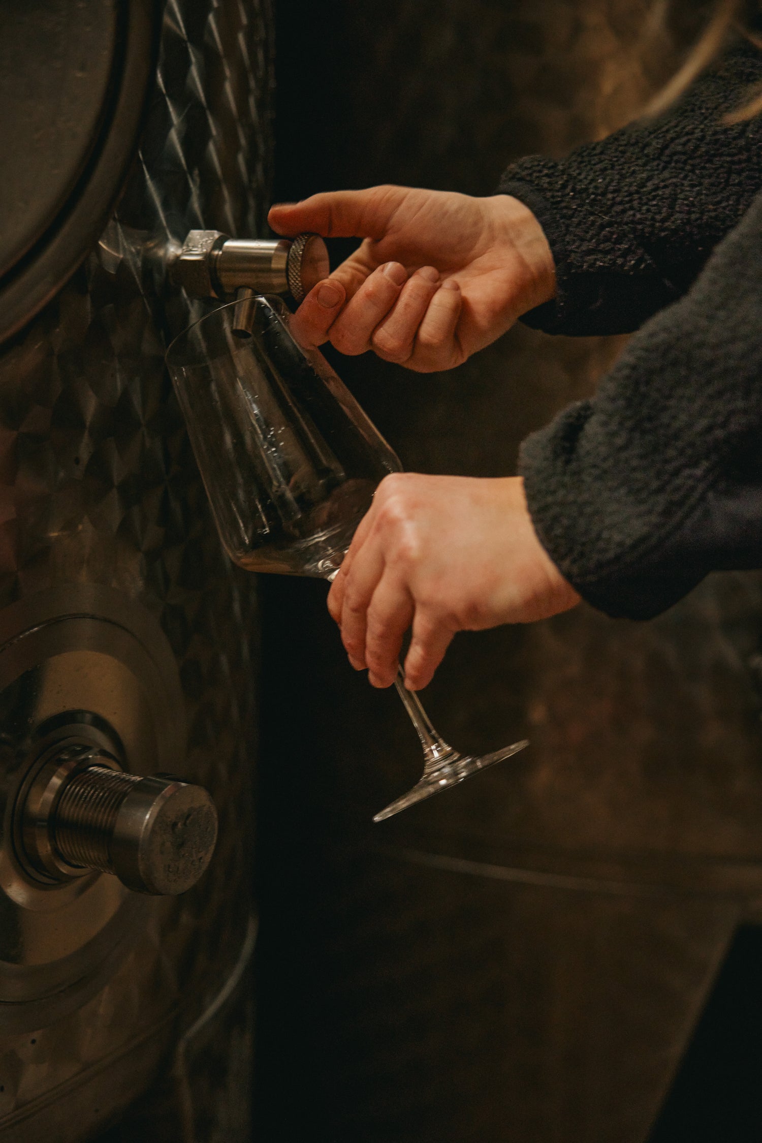 Ein Bildausschnitt von I AM NOT WINE Gründerin Franziska Treuer im Weinkeller. Sie hält mit der linken Hand ein Weinglas vor den Zapfhahn eines Weintanks. Mit der rechten Hand steuert sie den Zapfhahn, um etwas von dem Grundwein abzuzapfen.